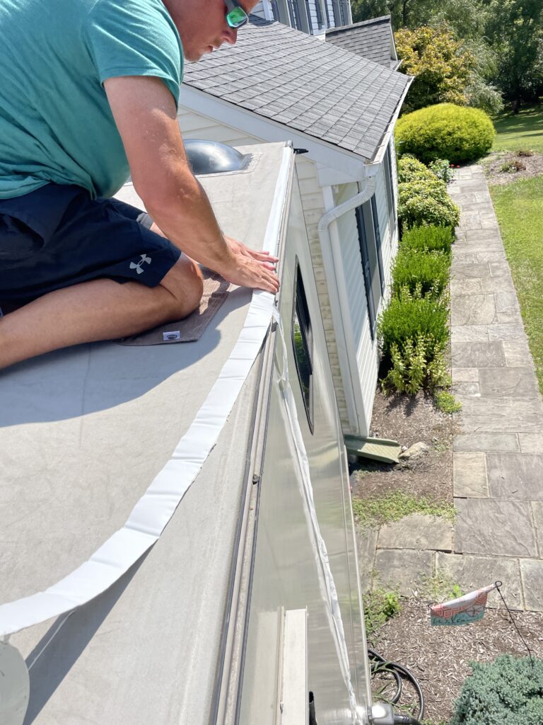 Man on roof of RV demonstrating how he is applying sealing tape to edge of roof line.